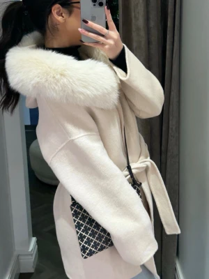 Alicia Coat Taupe med äkta päls  - Säljer en snygg off white  kappa med stor äkta päls runt kragen och knytskärp i midjan. Jackan har raka ärmar, stora fickor framtill och en oversized passform. Perfekt för dig som vill ha en trendig och mysig look under höst och vinter. Kappan består av 90% ULL och 10% kashmir, pälskragen består av 100% rävpäls. Använt jackan 4 gånger max, inga täcken på användning. 
