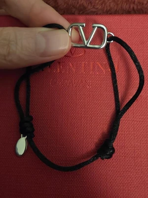 Valentino svart armband  - Snyggt armband från Valentino Garavani med svart justerbart band och ikonisk silvrig logga framtill. Metallbricka med gravyr och diskret design som passar till allt. Kommer med originalpåse och ask. Kan mötas i Stockholm 