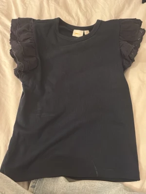 topp med volangärm - Säljer en marinblå topp från Gina Tricot med ribbad struktur och breda volangärmar i broderad bomull. Ärmarna har fina håldetaljer och vågig kant som ger en söt touch. Perfekt till jeans eller kjol för en trendig look.