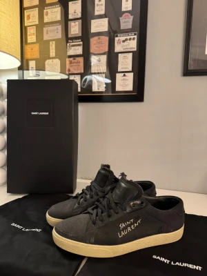 Svarta Saint Laurent sneakers (court classic) - Svarta sneakers från Saint Laurent med handskriven logga på sidan. Skorna har slitna detaljer, svart textil och läder, samt beige platt gummisula. Klassisk snörning och låg profil gör dem till ett statement i garderoben. Kommer med originalkartong och dustbags. Köpta på farfetch för cirka 6000. Storlek 39,5 men väldigt stora i storleken. Säljer dessa för 3500 men kan tänka mig att byta mot ett par andra skor.