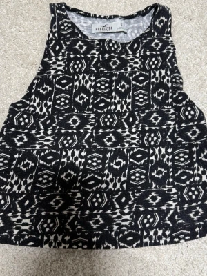 Svartvit mönstrad linnetopp Hollister - Säljer en svartvit linnetopp från Hollister i storlek S. Toppen har ett grafiskt mönster med tribal-inspirerade detaljer och är ärmlös med rund halsringning. Perfekt till sommaren och lätt att matcha med jeans eller shorts.