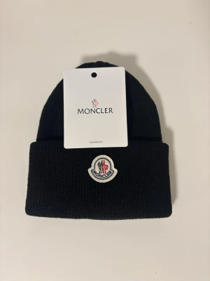 Svart Moncler mössa i ull - Snygg svart Moncler mössa med klassisk ribbstickad design och uppvikt kant. Framtill sitter Monclers ikoniska logotyp i vitt, rött och blått. Perfekt accessoar för kalla dagar och ger en clean look till din outfit.