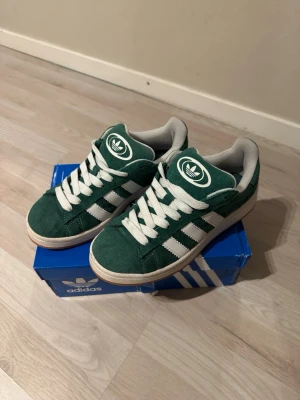 Adidas Campus gröna sneakers  - Säljer ett par gröna Adidas Campus sneakers i mocka med vita detaljer och klassiska tre ränder på sidan. Skorna har vit sula, vita skosnören och Adidas-logga på plös och häl. Perfekta för dig som gillar retrostil och streetwear. Använda några gånger i somras, annars har bara stått i lådan. 