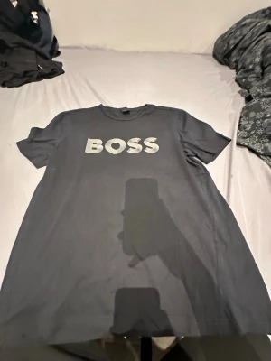 Svart BOSS t-shirt med tryck XL - Svart t-shirt från BOSS i storlek XL med stort vitt BOSS-tryck på bröstet. Klassisk rund hals och korta ärmar. Materialet är mjuk bomull som känns skönt mot huden. Perfekt för dig som gillar stilrena och tydliga loggor.