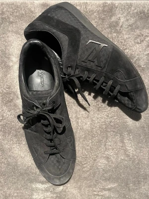 Svarta Louis Vuitton sneakers i mocka - Säljer ett par svarta Louis Vuitton sneakers med klassisk LV-logga på sidan. Skorna har snörning, rund tå och är tillverkade i en mix av mocka och textil. Snygg och stilren design med diskreta detaljer och LV-mönster på ovandelen. Perfekta för dig som gillar exklusiva sneakers.