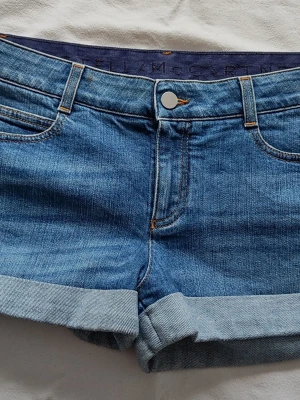 Blå jeansshorts från Stella McCartney, Strl 29 - Säljer ett par klassiska blå jeansshorts från Stella McCartney. De har upprullade benslut och snygga kontrastsömmar. Perfekta för sommaren och tillverkade i bomull. Stängs med knapp och dragkedja framtill.