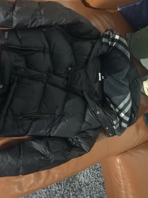 Svart pufferjacka från Burberry - Säljer en svart pufferjacka från Burberry med klassiskt rutigt foder i huvan och tydlig Burberry London England-patch på ärmen