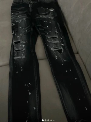 Svarta Dsquared2 jeans med slitningar - Säljer ett par svarta jeans från Dsquared2 med slim fit. Byxorna har slitna detaljer, hål och vita färgstänk för en riktigt edgy look. Klassisk femficksmodell med Dsquared2-logga på fickan och patch bak. Tillverkade i bomull.