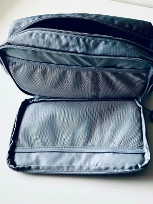 Grå sminkväska med meshfack - Rymlig necessär i grått tyg med flera smarta meshfack och dragkedjor. Många praktiska fack, se bilder. 28 x 18 x 10 cm. Perfekt för att organisera borstar, paletter och andra sminkprodukter. Praktiskt handtag på sidan och robust material som håller formen. Väskan har två stora huvudfack och flera mindre innerfickor.