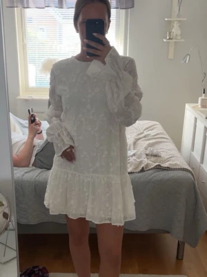 Vit blommig kortklänning från NA-KD - Populära studentklänningen!!!Supersöt vit kortklänning från NA-KD med blommigt broderi, volang nertill och långa puffiga ärmar. Klänningen har öppen rygg med knytband. Perfekt för sommarens alla tillfällen!