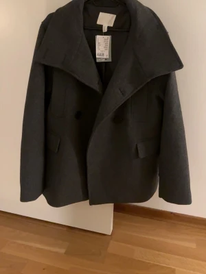 Mörkgrå skepparkavaj från H&M, S - Säljer en stilren mörkgrå skepparkavaj från H&M i storlek S. Jackan har bred krage, dubbelknäppning framtill och två fickor med lock. Materialet är ullblandning som ger en klassisk look och passar perfekt till höst och vinter.