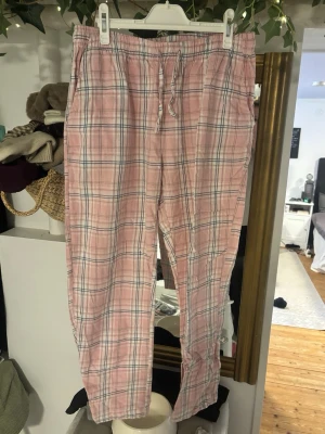 Rutig rosa byxa med snörning - Säljer en rutig byxa i ljusrosa toner med vita och mörkblå ränder. Västen har snörning i midjan för justerbar passform och är tillverkad i mjukt bomullstyg. Perfekt för dig som gillar avslappnad och trendig stil.