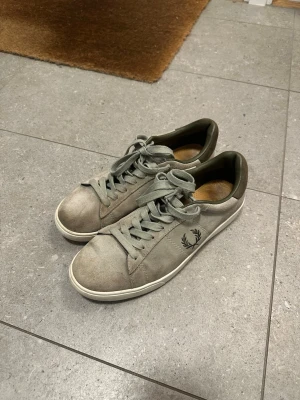 Fred Perry Skor - Ett par gröna Fred Perry sneakers i mocka, några smutsfläckar finns. En tydlig fläck finns på den högra skons tå.