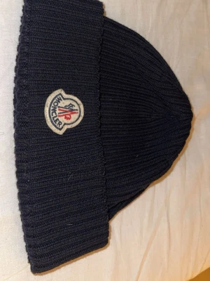 Svart ribbstickad mössa från Moncler - Säljer en svart ribbstickad mössa från Moncler med klassisk uppvikt kant och broderad logotyp framtill. Mössan är tillverkad i Italien och har en stilren, enkel design som passar perfekt till vintern.