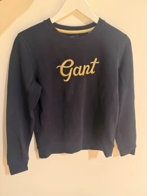 Svart sweatshirt från Gant - Mörkblå sweatshirt från Gant med guld broderad logga på bröstet. Klassisk rund halsringning och långa ärmar. Perfekt för dig som gillar stilrena och bekväma plagg med en touch av lyx. Materialet känns mjukt och skönt mot huden.