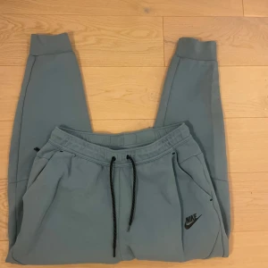  Nike tech fleece byxor - Säljer ett par ljusblå mjukisbyxor från Nike med svart logga på vänster ben. Byxorna har resår i midjan med svarta snören, muddar vid bensluten och praktiska fickor, varav en bakficka med dragkedja. Perfekta för chill eller träning. (Liten fläck på baksidan, se sista bild)