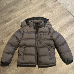 Grå dunjacka från Polo Ralph Lauren - Säljer en grå dunjacka från Polo Ralph Lauren med huva och flera dragkedjeförsedda fickor. Jackan har en puffig look, diskret logga på bröstet och är perfekt för kalla dagar. Klassisk design med lång ärm och rejäla muddar.