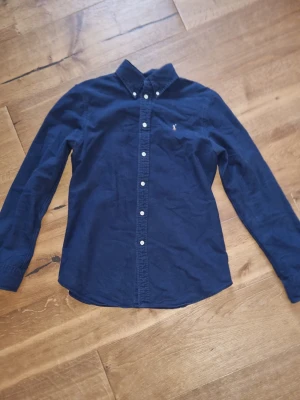 Mörkblå skjorta med knappar - Snygg mörkblå skjorta med klassisk krage och vita knappar hela vägen. Liten broderad logga på bröstet ger en clean look. Skjortan har lång ärm och är gjord i mjuk bomull, perfekt till jeans eller chinos. Passar s också