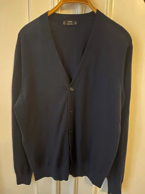 Mörkblå kofta i merinoull från Mango - Snygg mörkblå kofta från Mango i mjuk merinoull. Klassisk v-ringad modell med knappar framtill och långa ärmar. Perfekt att slänga över en t-shirt eller skjorta för en clean och stilren look. Storleken M är ganska true to size men kan även passa dem som bär S. Nypris 600kr.