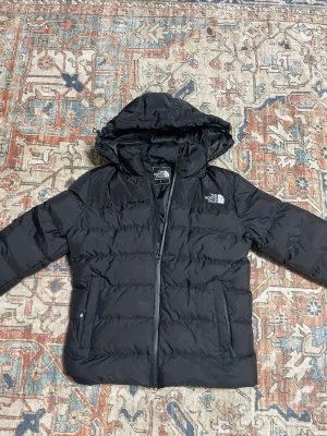 The North Face - Säljer en svart pufferjacka från The North Face i storlek S. Jackan har huva, dragkedja framtill och två sidofickor. Perfekt för kalla dagar och har en clean, stilren look med logga på bröstet och ryggen.
