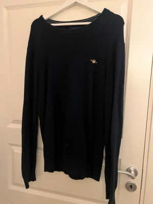 Mörkblå stickad tröja från GANT - Säljer en mörkblå stickad tröja från GANT i storlek L. Tröjan har rund halsringning med ribbade detaljer och en liten broderad GANT-logga på bröstet. Perfekt till höst och vinter för en clean och klassisk look.