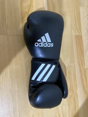 Svarta Adidas boxningshandskar - Säljer ett par svarta boxningshandskar från Adidas med klassiska vita ränder och logga. Snygg och clean design som passar perfekt till träningen.