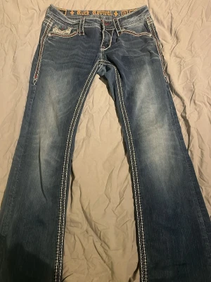 Rock Revival blå bootcut jeans - Säljer ett par blå jeans från Rock Revival med tydliga kontrastsömmar och broderade detaljer på bakfickorna. Jeansen har bootcut-modell och låg midja, med slitningar och snygg tvätt. Materialet är klassisk jeansbomull och passformen är normal. Storleken är 27 men passar på mig som är 28/29. Denna model säljs ej längre. 