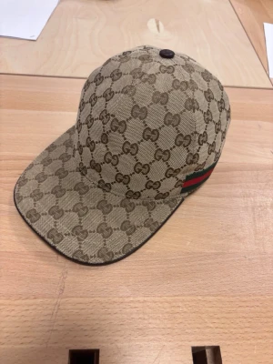  beige keps med GG-mönster - Säljer en beige keps från Gucci med det klassiska GG-mönstret över hela kepsen. Kepsen har en justerbar rem baktill och en snygg detalj med grönt och rött band på sidan. Materialet är canvas och detaljerna är i brunt.