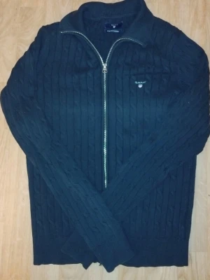 Mörkblå kabelstickad kofta från GANT - Säljer en mörkblå kabelstickad kofta från GANT med dragkedja framtill och diskret logga på bröstet. Tillverkad i stretchig bomull för extra komfort. Perfekt till lager på lager och har en klassisk, stilren look.