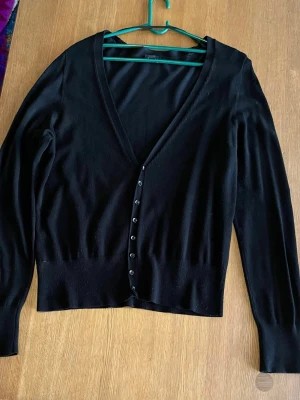 Svart kofta med v-ringning från Lindex - Säljer en svart kofta från Lindex i storlek L. Koftan har djup v-ringning, långa ärmar och knappar framtill. Perfekt att slänga över en topp eller klänning för en enkel men snygg look.