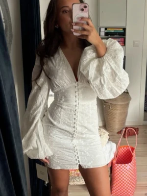 Vit kort klänning med puffärmar - Superfin vit kort klänning med broderade blommor, djup v-ringning och stora puffärmar. Klänningen har knappar framtill och markerad midja som ger en snygg siluett. Perfekt för sommarens alla tillfällen!