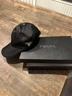 Prada Keps - ALDRIG ANVÄND. Bilderna visar en svart keps från Prada med guldig detalj. 