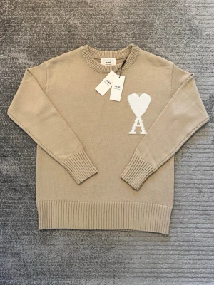 Beige stickad tröja från Ami - Säljer en beige stickad tröja från Ami Alexandre Mattiussi med ikoniskt vitt hjärta och A på bröstet. Tröjan har rund hals, ribbade muddar och nederkant samt lång ärm. Perfekt för dig som gillar stilrena och trendiga plagg.