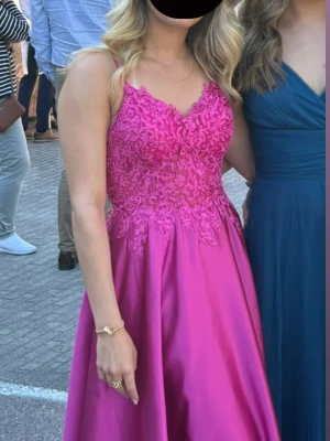 Balklänning med spets - Säljer en magentafärgad långklänning med spetsdetaljer och tunna axelband. Kjolen är slät och faller snyggt ner till golvet. Perfekt för bal eller student. Klänningen har v-ringning och vackert broderat mönster.