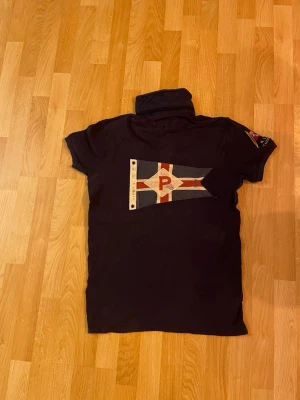 Svart pikétröja från Polo Ralph Lauren - Svart pikétröja från Polo Ralph Lauren med vit diagonal rand och röd logga på bröstet. Baksidan har ett stort flaggtryck och detaljer på ärmen. Klassisk krage och korta ärmar, tillverkad i bomull.passar 167-174,
