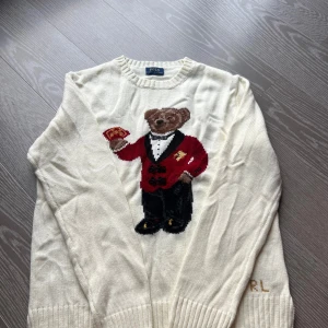 Vit stickad tröja Polo Bear Ralph Lauren - Vit stickad tröja från Polo Ralph Lauren med ikoniska Polo Bear-motivet framtill. Björnen har röd kavaj och håller spelkort. RL-broderi i guld på ärmen. Klassisk rund hals och ribbade muddar. Perfekt statement-piece för dig som gillar preppy stil. Längd 63 cm bredd 49 cm. S/Xs