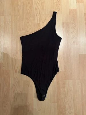 Svart one shoulder body från Hollister - Svart ribbad body från Hollister med one shoulder-design och tryckknappar i grenen. Snygg och enkel modell som är lätt att styla till olika outfits. Tillverkad i mjukt material som sitter skönt mot kroppen.