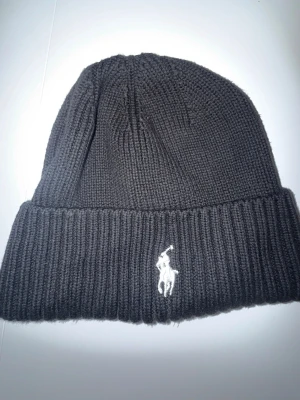 Ralphlauren mössa - Säljer en svart stickad mössa från Polo Ralph Lauren med klassisk ribbad kant och den ikoniska vita loggan broderad framtill. Perfekt accessoar för kyliga dagar och enkel att matcha med olika outfits.