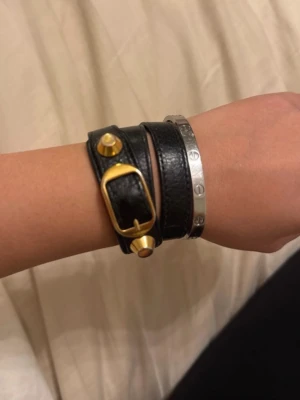 Svart läderarmband från Balenciaga - Säljer ett svart armband i skinn med dubbla remmar, guldfärgat spänne och nitar. Snygg accessoar som ger en edgy vibe till din outfit. Perfekt att matcha med andra smycken för en cool lager-på-lager-look. Från Balenciaga - köpt på Plick har serienummer i, ej kvitto