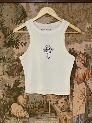 Vit Lady Gaga Tour linnetopp - Säljer en vit linnetopp från Lady Gaga Tour 2025/2026 med tryck av ett kors och texten 'Mayhem' framtill samt turnétryck på ryggen. Toppen är ärmlös och croppad, tillverkad i en mjukt tyg. 