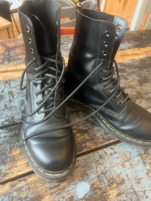Dr. Martens 10-hål - Klassiska svarta Dr. Martens boots i skinn med högt skaft och gul söm runt sulan. Skorna har snörning hela vägen upp och en robust, platt sula. 