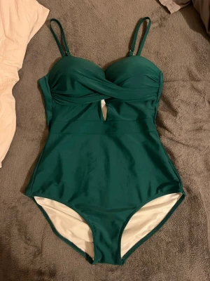 Grön baddräkt med cut-out detalj - Snygg grön baddräkt med justerbara axelband och en liten cut-out detalj framtill. Baddräkten har vadderade kupor och en enkel, stilren design utan mönster. Perfekt för strand eller poolhäng!