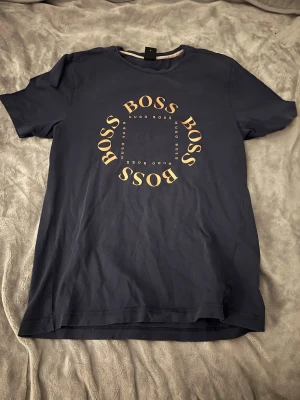 Mörkblå Hugo Boss t-shirt med tryck - Snygg mörkblå t-shirt från Hugo Boss med rund hals och korta ärmar. Framtill finns ett stort tryck med texten 'BOSS' i en cirkel och mindre 'Hugo Boss' i mitten. Tillverkad i mjuk bomull för skön känsla och stilren look.
