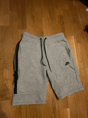 Grå Nike shorts  - Säljer ett par gråa Nike shorts med svart logga på vänster ben. De har snörning i midjan och en svart dragkedja längs sidan. Tillverkade i mjuk bomullsblandning, perfekta för chill eller träning.