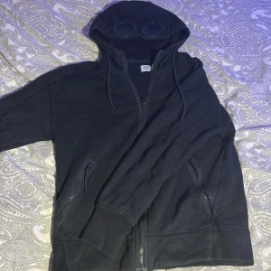 Svart hoodie från C.P. Company - Svart hoodie från C.P. Company med ikoniska goggles i huvan. Tröjan har dragkedja framtill, två fickor med dragkedja och ribbade muddar. Perfekt för dig som gillar streetwear och vill sticka ut med en unik detalj på huvan.