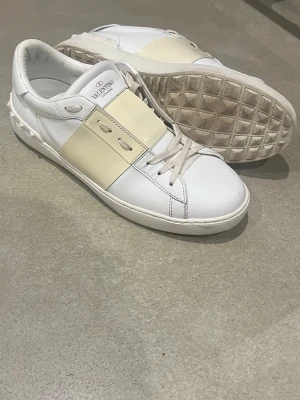 Valentino Garavani vita sneakers - Säljer ett par vita Valentino Garavani sneakers med ikoniska nitar på hälen och breda beigea paneler på sidorna. Skorna har snörning, platt sula och är tillverkade i skinn. Perfekta för dig som vill ha en clean men ändå lyxig look.