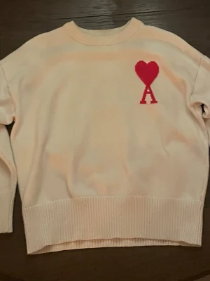 Vit stickad tröja från Ami - Säljer en vit stickad tröja från Ami Alexandre Mattiussi med röd hjärtdetalj och bokstaven A på bröstet. Tröjan har rund halsringning, ribbade muddar och lång ärm. Perfekt för dig som vill ha en clean men ändå unik look.