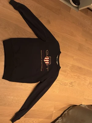 Mörkblå sweatshirt från Gant - Säljer en mörkblå sweatshirt från Gant med klassiskt tryck och logga framtill i rött, vitt och guld. Tröjan har rund halsringning och långa ärmar. Perfekt till chill dagar och enkel att matcha med jeans eller sweatpants.