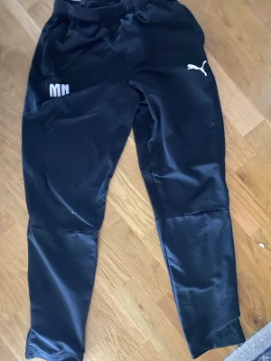 Svarta träningsbyxor från Puma - Säljer ett par svarta träningsbyxor från Puma med vit logga på låret och elastisk midja. Byxorna har smal passform och är gjorda i ett glansigt syntetmaterial som andas bra. Perfekta för träning eller chill. Initialerna MN broderade på ena sidan.