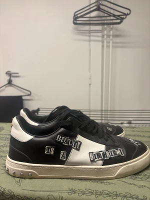 Valentino Garavani svartvita sneakers - Unika svartvita sneakers från Valentino Garavani med coola textpatches, stjärnor broderade på hälen och nitar runt sulan. Skorna har snörning och är tillverkade i skinn. Sulan har ett camomönster undertill och detaljerna gör dem riktigt statement.